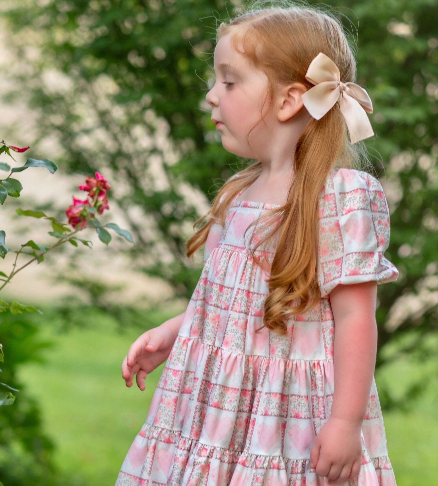 little girl playing in an outdoor scene with lots of greenery, wearing Puff sleeve dress with a soft white background, dress has a block print reminiscent of quilting with faint watercolor looking pink hearts in the center on some blocks, and a floral print in other blocks. The skirt of the dress is sewn into three separate tiers, and the waist is in the empire style with a square neckline. 