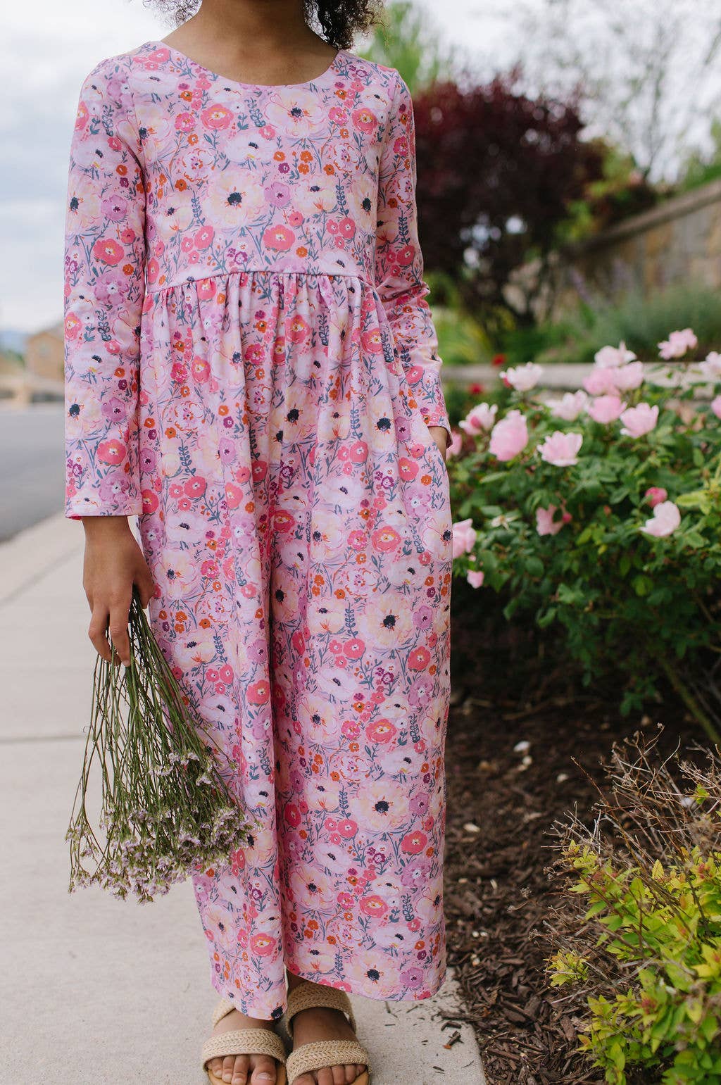 Person wearing a pink floral dress standing outdoors with flowers and greenery.