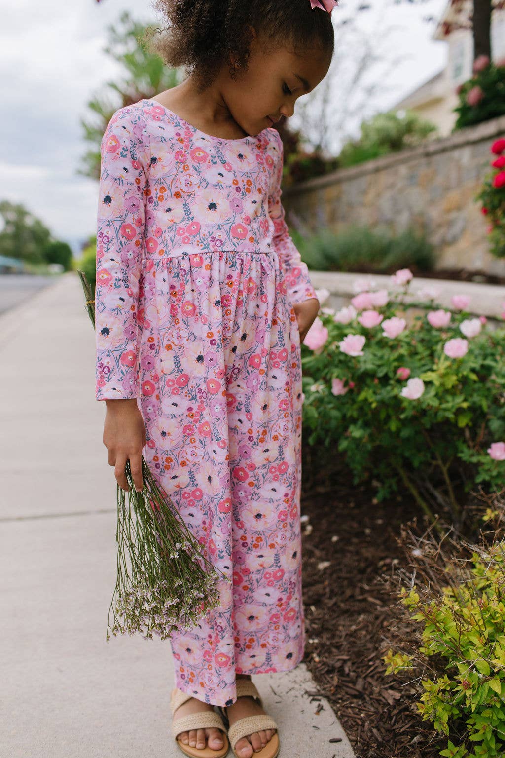 Child in a floral dress standing outdoors next to a garden.
