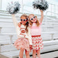 Two young girls in festive outfits with pom-poms and sunglasses on a bleacher background.