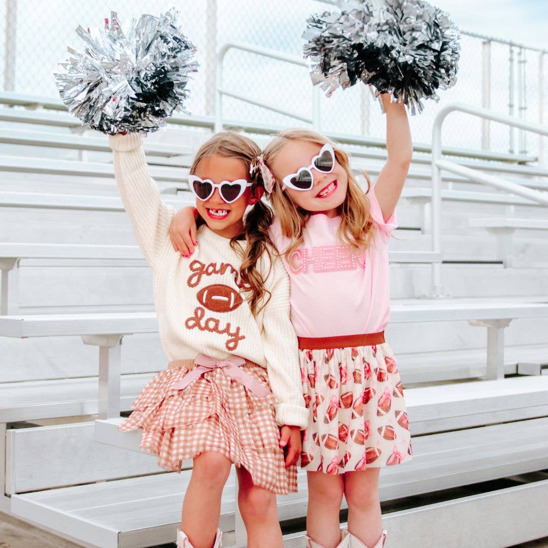 Two young girls in festive outfits with pom-poms and sunglasses on a bleacher background.