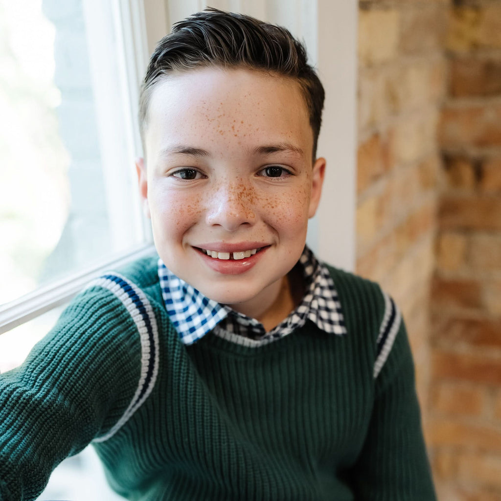 Young boy wearing a green sweater with a checkered shirt underneath, sitting indoors.
